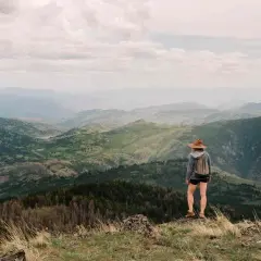 Una excursionista con sombrero admira las majestuosas montañas de Madrid, un paisaje verde y brumoso bajo un cielo nublado.