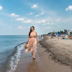 Kobieta w kwiecistej sukience spaceruje brzegiem morza. W tle plaża pełna ludzi, parawanów i domków – typowe sarbinowo atrakcje.