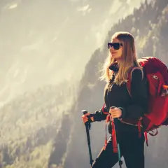 Kobieta w okularach przeciwsłonecznych i z plecakiem, z kijkami trekkingowymi, pokazuje, jak się ubrać w góry wiosną.