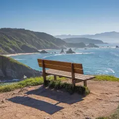 Un banco vacío invita a contemplar el mar azul desde el mirador de Coitelo, con acantilados verdes y rocas emergiendo del agua.