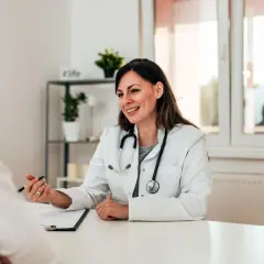 Medico sorridente parla con paziente, illustrando un punto con la penna. La scena evidenzia una buona comunicazione medico-paziente.