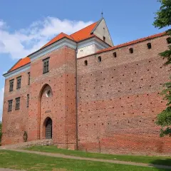 Zamek w Łęczycy, ceglana twierdza z czerwonej cegły, z charakterystycznym łukowym wejściem i oknami, otoczona zielenią.