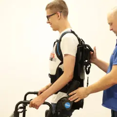 Mężczyzna w okularach ćwiczy chód w egzoszkielecie, wspierany przez terapeutę. Trójnóg rehabilitacyjny pomaga w pionizacji.