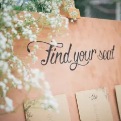 Panneau en bois avec l'écriture "Find your seat" et des fleurs. Des cartes de placement sont fixées en dessous.
