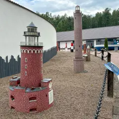 Miniaturowa latarnia morska z czerwonej cegły, obok wyższa, kamienna. W tle budynek i łódź.