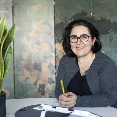 Frau mit Brille lächelt, hält Stift. Im Hintergrund Weltkarte. Sie berät zu Lohnsteuerhilfeverein Kosten.