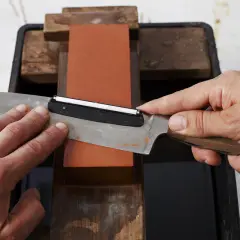 Händer slipar en kniv mot en orange sten. En vass kniv är viktig för matlagning.