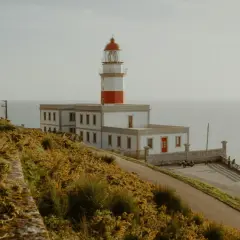 El faro de Baiona se alza majestuoso sobre un acantilado cubierto de vegetación dorada, vigilando el mar.