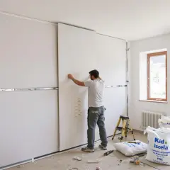 Un ouvrier installe des panneaux blancs sur une structure métallique, préparant un faux plafond. L'épaisseur du faux plafond est visible sur une note collée au panneau.