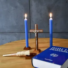 Dwie niebieskie świece, krzyż, kropidło i Biblia Jerozolimska na drewnianym stole. Symbolika religijna, jakby przygotowanie do kolędy.