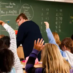 Schüler heben in einer finnischen Schule die Hände. Eine Lehrerin steht vor einer Tafel mit mathematischen Aufgaben.