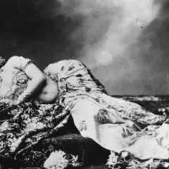 Mata Hari, bailarina exótica, yace recostada sobre telas estampadas, con un tocado adornado y mirada cautivadora.