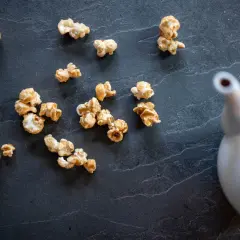 Pop corn caramel éparpillé sur une surface sombre, avec une théière blanche en arrière-plan.