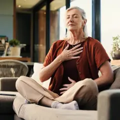 Äldre kvinna sitter på en soffa och fokuserar på sin andning för att hantera anxiety svenska. Hennes händer vilar på bröstet och magen.