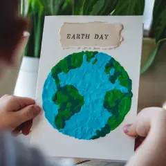 Dziecko trzyma kartkę z pracą plastyczną przedstawiającą planetę Ziemię z plasteliny. Napis "EARTH DAY" na górze.