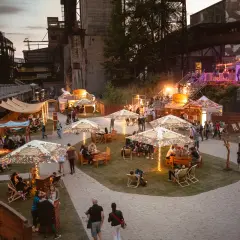 Besucher genießen das Ostrava Festival in einer ehemaligen Industrieanlage. Stände, Sitzbereiche und Lichter schaffen eine festliche Atmosphäre.