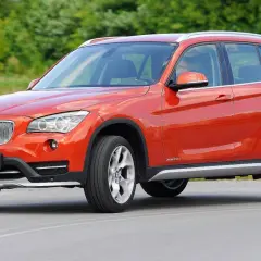 Pomarańczowe BMW E84 X1 z polskimi tablicami rejestracyjnymi jedzie po drodze, otoczone zielenią.