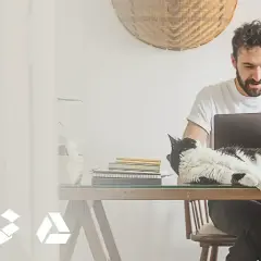 Mężczyzna pracuje przy laptopie, a kot leży na biurku. Ikony chmury, pudełka i trójkąta symbolizują online photo storage options.