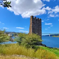 Torre medieval junto a un río azul en Catoira, Galicia. Un puente cruza el agua al fondo.