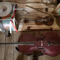 Instrument ludowy podobny do wiolonczeli, skrzypce i bębenek leżą na drewnianej podłodze.