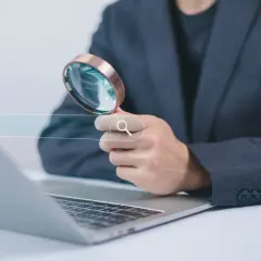 Osoba w garniturze trzyma lupę nad polem wyszukiwania na tle laptopa.