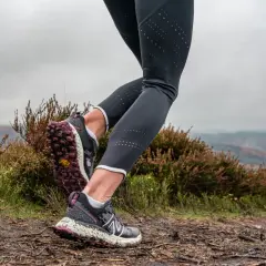 Gambe di una runner su un sentiero fangoso, con erica e colline sullo sfondo. Perfetto per chi vuole iniziare il trail running.