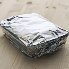 Ein rechteckiges Paket, in glänzende Alufolie gewickelt, liegt auf einem Holztisch. Die Frage, welche Seite der Alufolie giftig ist, bleibt unbeantwortet.