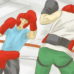 Two boxers in a ring, one in a blue shirt and red headgear, the other in a white shirt and grey cap, demonstrating how to box.