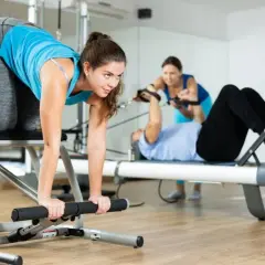 Frauen trainieren im Studio. Eine macht eine Übung auf einem Gerät, während andere im Hintergrund an Pilates-Geräten arbeiten. Das ist Zirkeltraining.