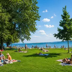 Sonniger Tag im Strandbad Bodensee. Menschen entspannen auf Decken im Gras, baden im See und genießen die Aussicht.