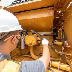 Pracownik w kasku i kamizelce konserwuje żółte urządzenia budowlane, sprawdzając przewody hydrauliczne.