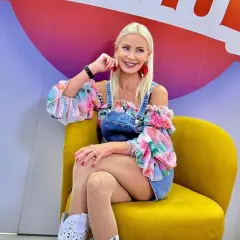 Uśmiechnięta Elwira Mejka w kwiecistej bluzce i jeansowym ogrodniczku, siedzi na żółtym fotelu.