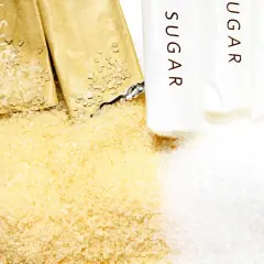 Zbliżenie na rozsypany cukier trzcinowy i biały, obok otwartych saszetek z napisem "SUGAR".