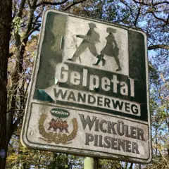 Schild am wanderparkplatz Gelpetal weist den Weg. Silhouetten von Wanderern und Bierwerbung.