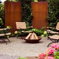 Moderne, schlichte Gartengestaltung mit vier Holzstühlen um eine Feuerstelle. Rostige Metallwände und üppiges Grün schaffen eine gemütliche Atmosphäre.