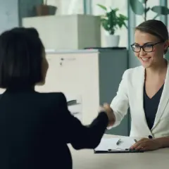 Zwei Frauen schütteln sich die Hände. Eine Frau im weißen Blazer lächelt, während sie eine Bewerbung für personalentwicklung jobs prüft.