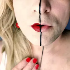 Un volto diviso a metà, con capelli biondi e barba, labbra rosse e una linea nera. Un'espressione di una persona non binaria che abbraccia la propria identità.
