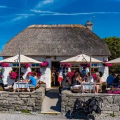 Café pittoresque avec toit de chaume, parasols et fleurs. Des gens dégustent un repas en plein air, peut-être un délicieux aran poisson.