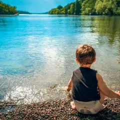 Un bambino seduto sulla riva di un lago, godendosi un tranquillo week end con bambini. L'acqua limpida riflette il cielo azzurro e gli alberi verdi.