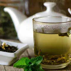 Dampfender grüner Tee in einer Tasse, daneben eine Schale mit Teeblättern und Minze. Welcher Tee ist antibakteriell? Grüner Tee ist bekannt für seine gesundheitlichen Vorteile.