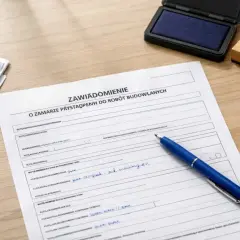 Formularz "Zawiadomienie o zamiarze przystąpienia do robót budowlanych" leży na biurku, gotowy do wypełnienia. Obok stos dokumentów i niebieski długopis.