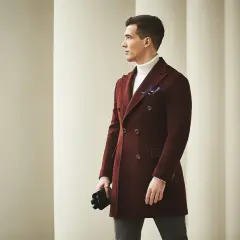 Elegancki mężczyzna w bordowej budrysówce i białym golfie, z czarnymi rękawiczkami w dłoni, pozuje na tle kolumn.
