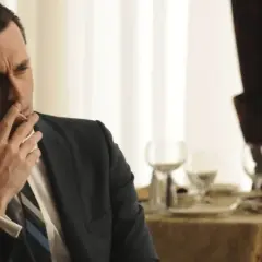 Jon Hamm, znany z filmów, seriali i programów, w eleganckim garniturze pali papierosa przy stole.