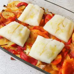 Cuatro porciones de bacalao al horno reposan sobre patatas y pimientos asados en una fuente de vidrio.