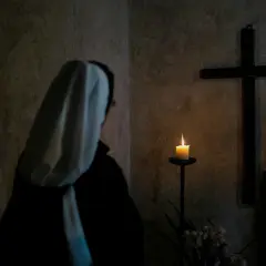 Zakonnica w białym habicie modli się przed krzyżem i płonącą świecą.