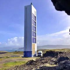 El faro de Punta Frouxeira se alza imponente sobre la costa, con un cielo azul y nubes blancas. Una furgoneta blanca descansa a su lado.