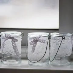 Drei hübsch gestaltete Windlichter aus Glas mit zarter Spitze und Schleifen.