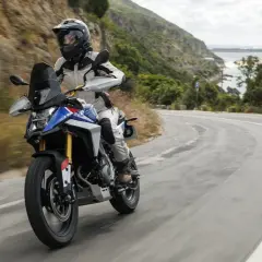 Motociclista en una maxi trail recorre una carretera sinuosa con vistas al mar y montañas verdes.