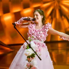 Sanah w kwiecistej sukni, z mikrofonem w dłoni, śpiewa na scenie. Jej **wzrost** i talent zachwycają publiczność.
