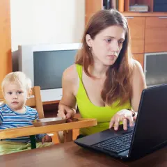 Matka z dzieckiem przy stole, zastanawia się, ile trwa urlop macierzyński, pracując na laptopie.